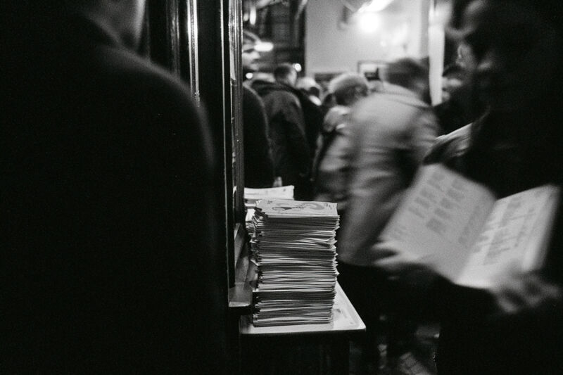 Schwarzweißfoto: ein Stapel Programmflyer liegt auf einer Ablage am Eingang, dahinter unscharf die wartenden Gäste
