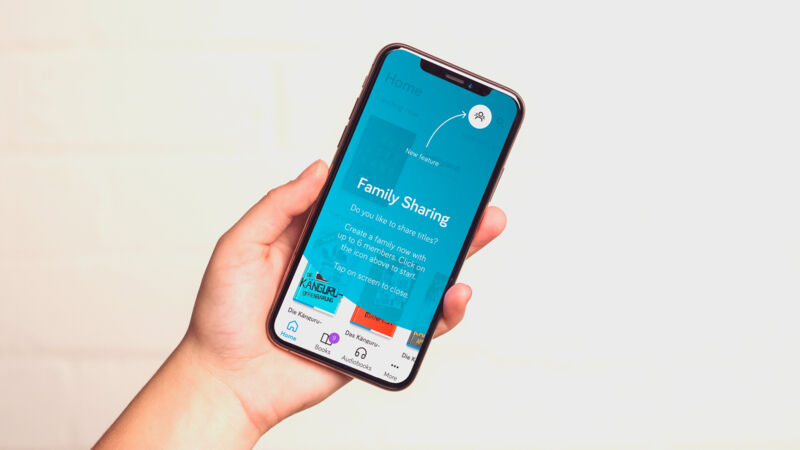 Eine Hand hält ein iPhone: über dem Startbildschirm der tolino App liegt der türkise Family-Sharing-Hinweis mit der Frage „Do you like to share titles?“ und der Aufforderung, eine Familie mit bis zu sechs Mitgliedern anzulegen. Ein geschwungener Pfeil zeigt auf das neue Familien-Icon in der Kopfleiste, darunter die Leseliste und die Tab-Leiste der App.